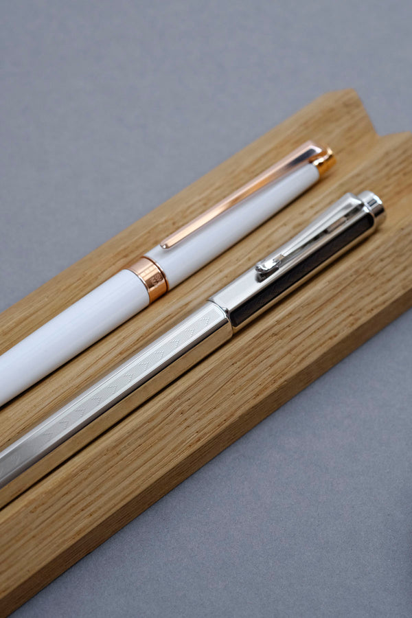 White Caran d’Ache Léman Slim fountain pen and Ecridor Chevron fountain pen on an oak stand.