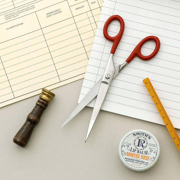 Steel scissors with red handles and the Penco brand name engraved on them, set against a lined notebook background.