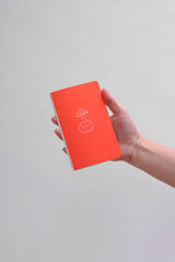 A small notebook in a vivid orange color with a white illustration, held in a hand.